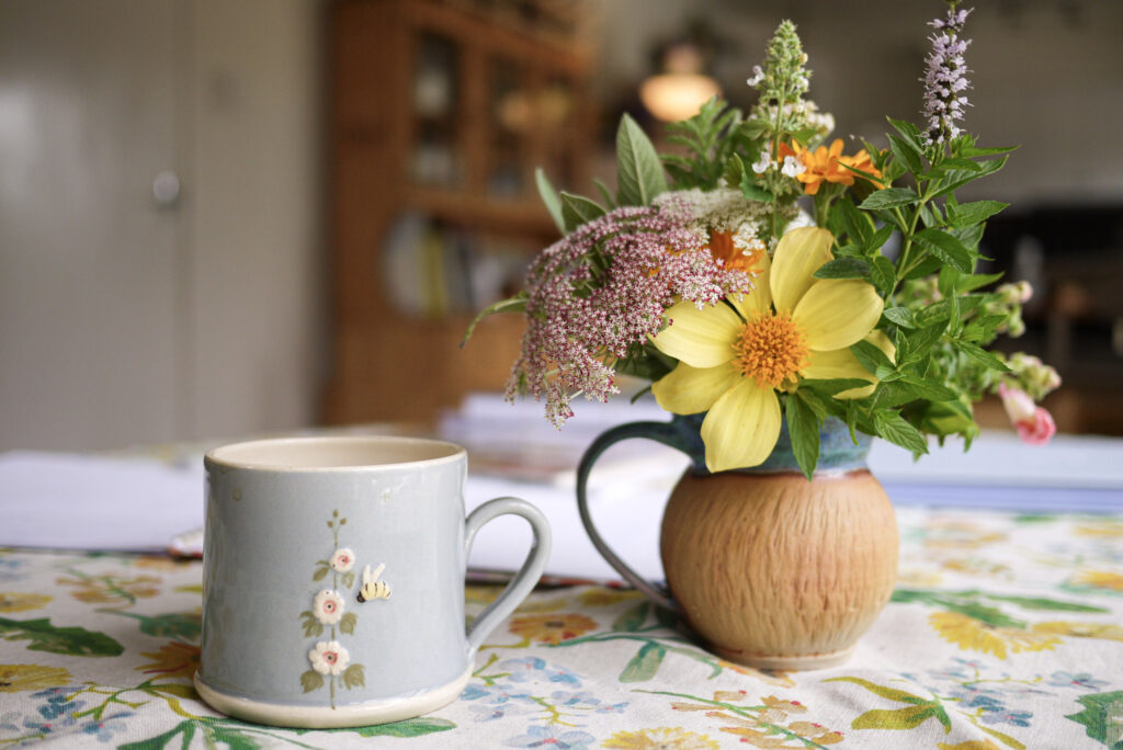 A ceramic teacup with a hollyhock and honey bee next to a small vase of homegrown flowers