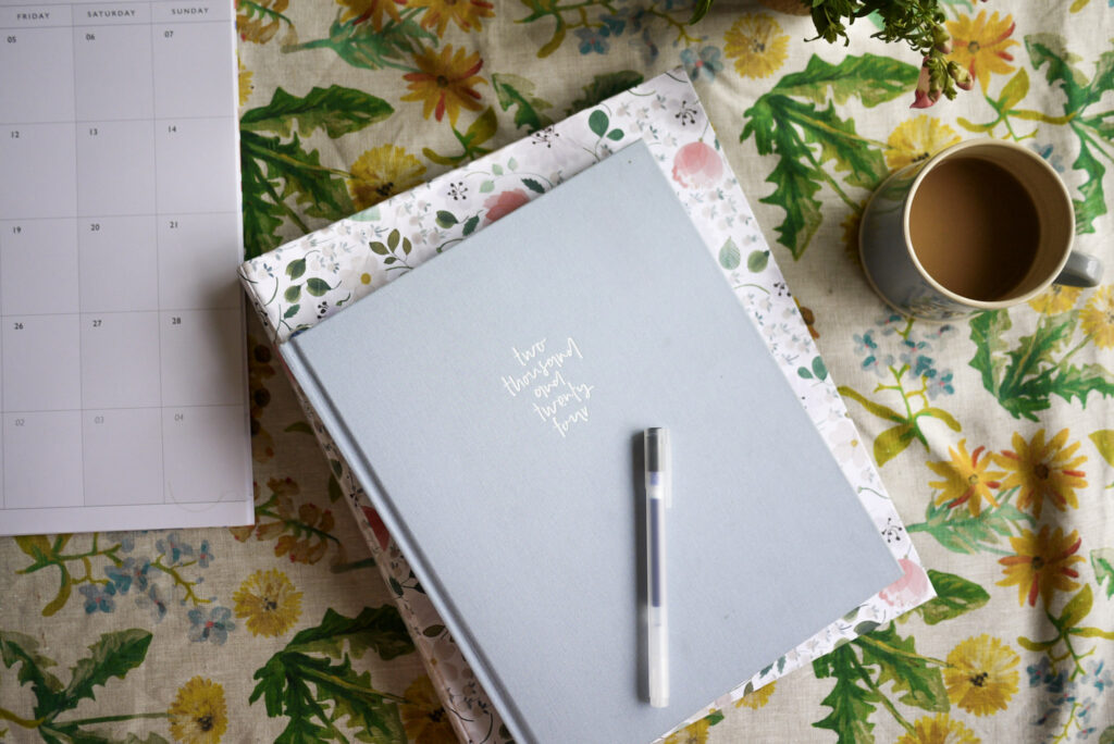 A paper calendar, home management binder, diary and cup of tea on a table with a dandelion linen table cloth